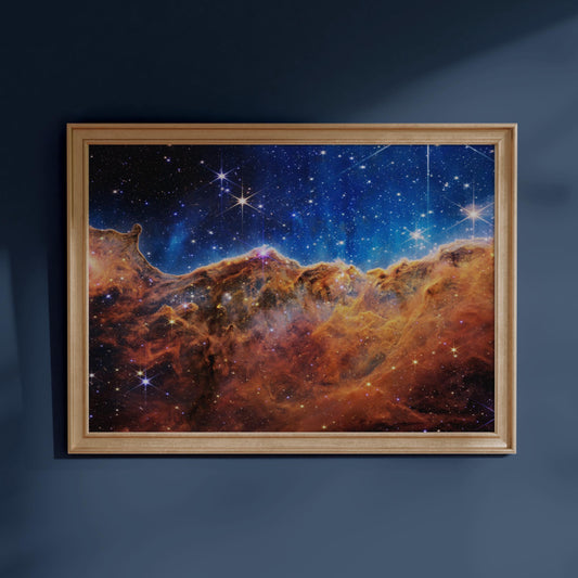 The image is a framed painting depicting a stunning nebula, a vast cloud of gas and dust in space, illuminated by the light of distant stars. The nebula is a vibrant mix of orange, red, and yellow hues, with numerous bright stars scattered throughout the scene. The painting is hung on a blue wall, creating a striking contrast between the celestial artwork and the surrounding environment.