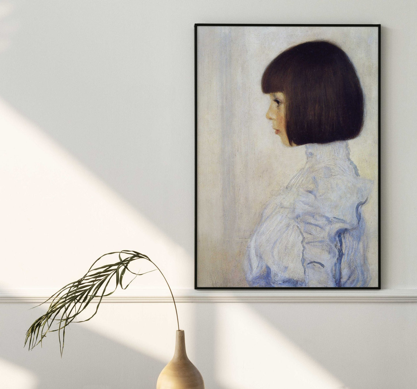 a framed portrait of a young girl with short dark hair, wearing a white blouse, hanging on a white wall. Next to the portrait is a small vase with a plant, creating a serene and elegant atmosphere.