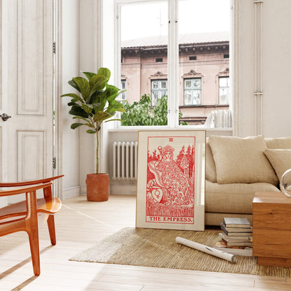a living room with a large window overlooking a building. The room is furnished with a couch, a chair, and a potted plant. A large framed tarot card, specifically "The Empress" card, is prominently displayed on the floor in front of the couch.